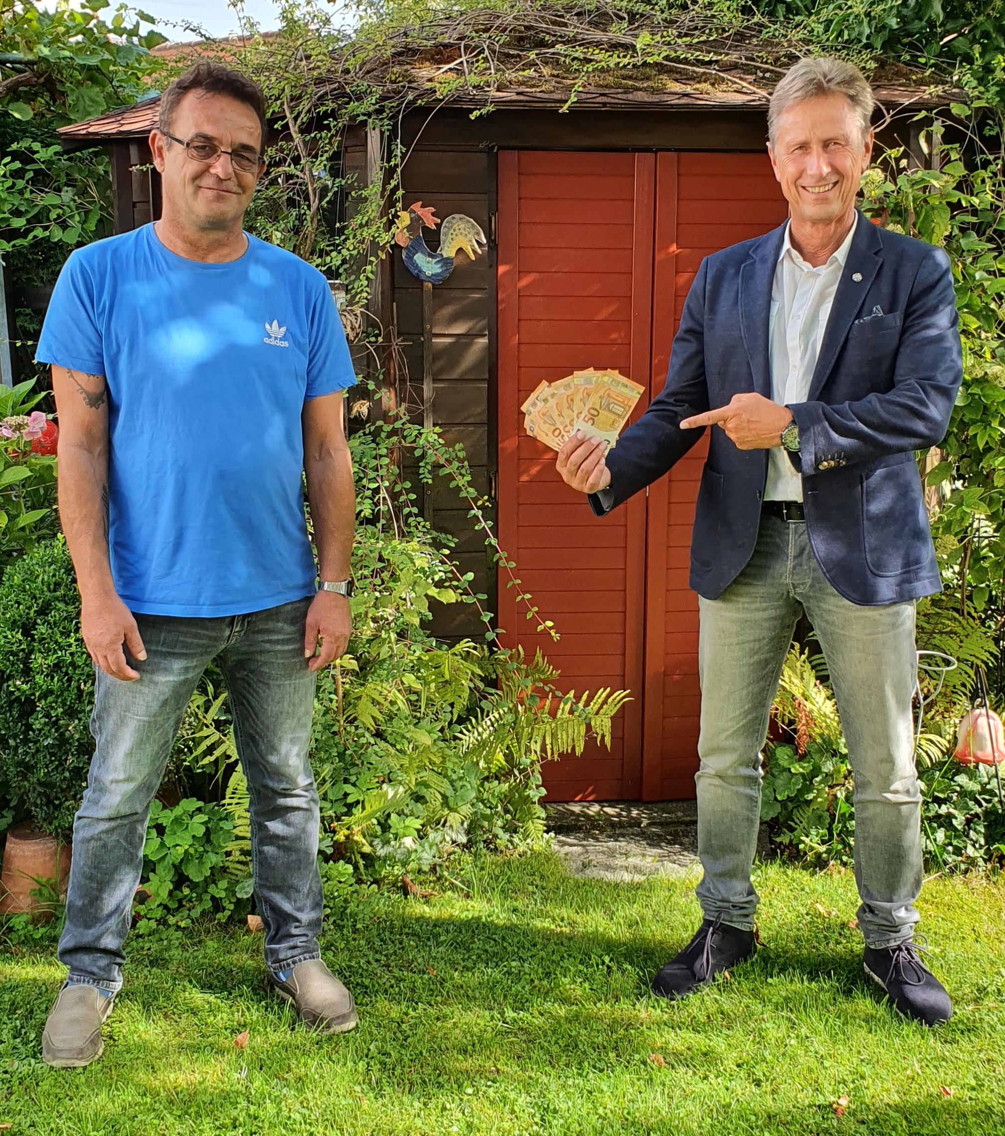 Zwei Männer vor Gartenschuppen, einer zeigt auf Geld in seiner Hand