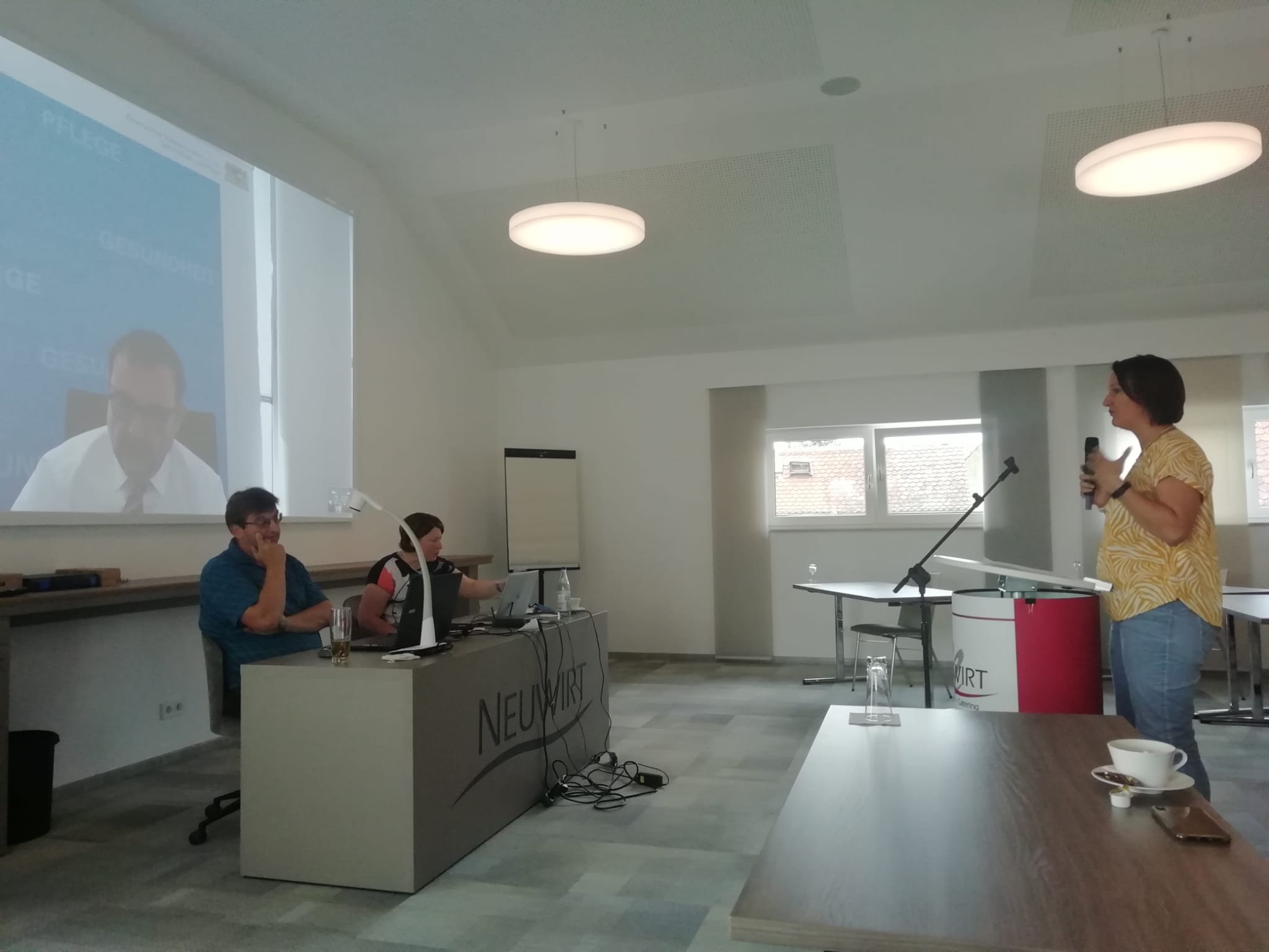 Leinwand mit Klaus Holetschek, davor Mann und Frau an einem Podium, rechts Frau an Mikrofonständer