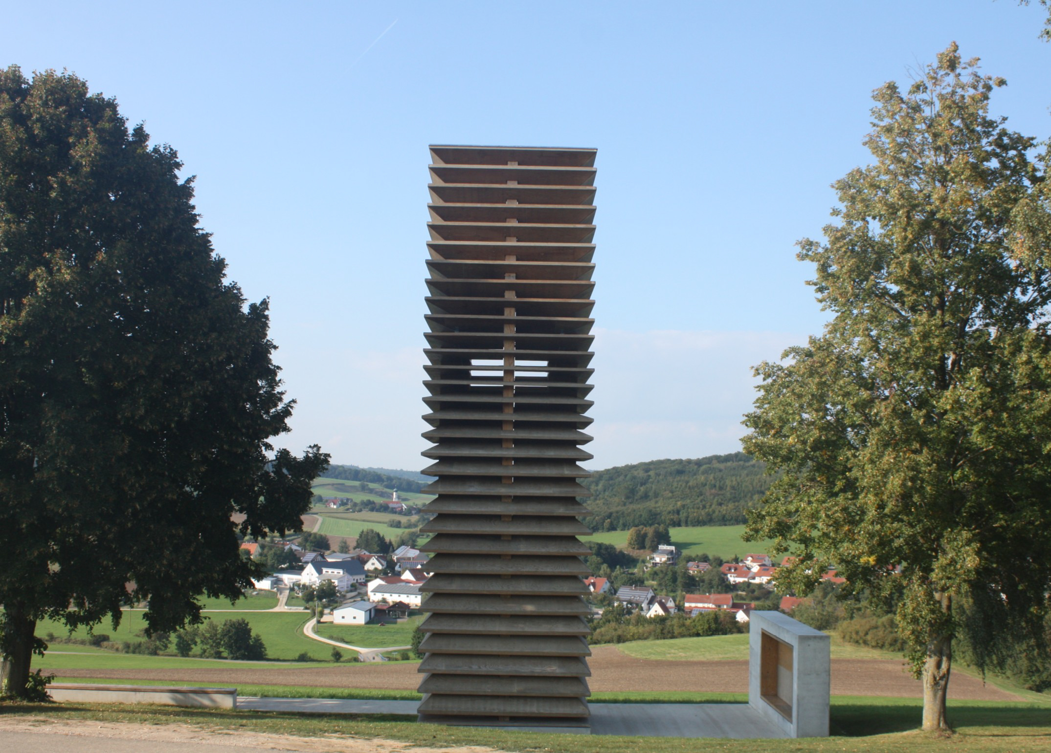 Turm aus Holzplatten zwischen Bäumen, im Hintergrund Häuser