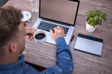 Mann mit Kaffeetasse sitzt am Tisch vor einem Laptop