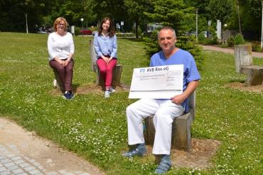 Drei Personen sitzen auf Wiese, Mann mit Scheck