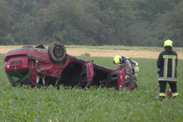 Roter Pkw liegt auf dem Dach in einem Feld, dabei zwei Feuerwehrleute