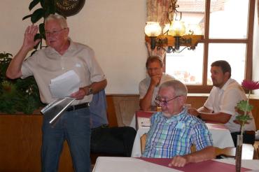 Vier Männer im Wirtshaus, drei sitzend, einer stehend, eine Rede haltend