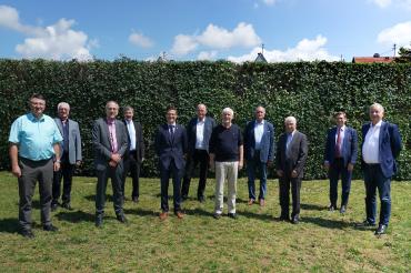 Mehrere Herren stehen in Anzug auf Wiese vor Hecke, mit Sicherheitsabstand