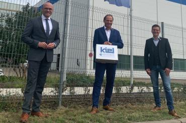 Drei Männer vor zwei Flaggen, dahinter Firmengebäude