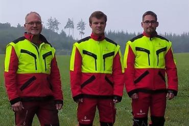 Drei Männer mit gelb-roter Warnkleidung auf Wiese vor Wald
