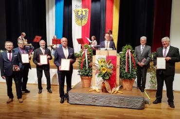 Sieben Herren im Anzug mit Urkunden vor Bühne, dahinter Flaggen