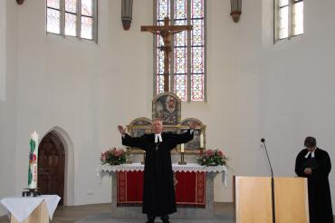 Pfarrer im Talar steht in Kirche vor Altar, Arme ausgebreitet