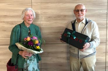 Ältere Dame und älterer Herr mit Blumenstrauß bzw. Geschenkschachtel vor Holzwand