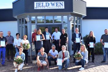17 Personen mit Urkunden bzw. Blumensträußen stehen bzw. knien vor JELD-WEN-Eingang