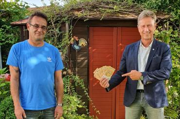 Zwei Männer vor Gartenschuppen, einer zeigt auf Geld in seiner Hand