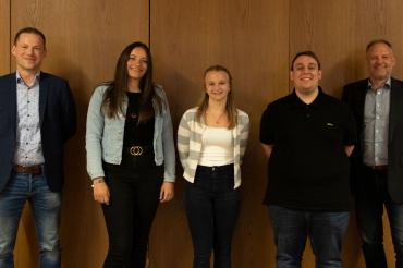 Zwei Frauen und drei Männer stehen vor einer Holzwand