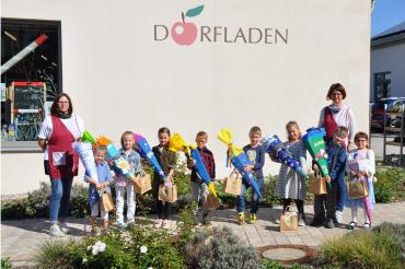 Das Bild zeigt Marion Dopfer (links) und Monika Schuster (rechts) zusammen mit den beschenkten Erstklässlerinnen und Erstklässlern vor dem Dorfladen in Rögling.