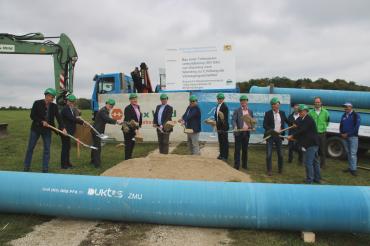 13 Herren beim Spatenstich, davor Wasserleitungsrohr