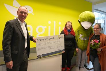 Drei Personen und ein Maskottchen mit Scheck vor agilis-Logo