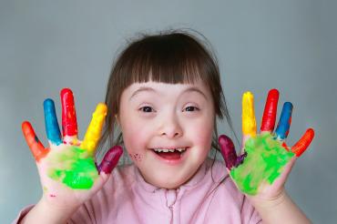 Das Bild zeigt ein Kind mit Trisomie 21, welches die Hände voller Farbe hat. 