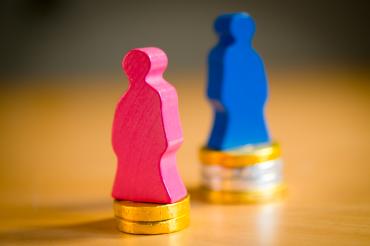 Pinke Holzfigur auf kleinem Münzstapel, blaue Figur auf größerem Münzstapel