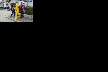 Gelbe Holzfigur auf Gehweg, daneben ein Junge und ein Mädchen, Gesichter unkenntlich gemacht