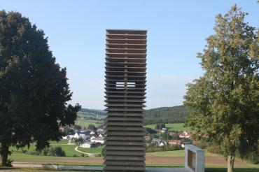 Turm aus Holzplatten zwischen Bäumen, im Hintergrund Häuser