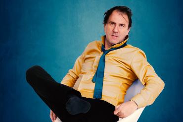 Mann im gelben Hemd mit blauer Krawatte sitzt vor blauem Hintergrund