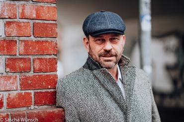 Michel Guillaume mit Mantel und Mütze an Backsteinmauer