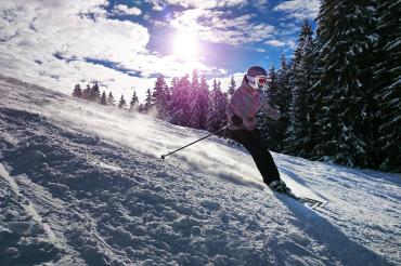 Skifahrerin fährt Berg hinunter, dahinter Bäume und tiefstehende Sonne