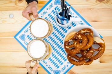 Zu sehen sind zwei Bierkrüge, welche von zwei Händen zum Anstoßen gehalten werden, daneben steht ein Teller mit Brezeln. 