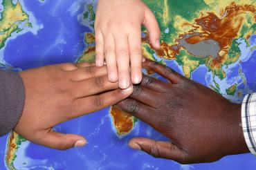 Zu sehen sind drei Hände mit unterschiedlichen Hautfarben, die auf einer Weltkarte liegen. 