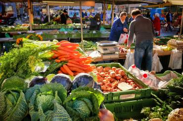Zu sehen ist ein Marktstand mit Gemüse, bei dem gerade jemand einkauft. 