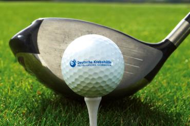 Zu sehen ist ein Golfball mit der Aufgschrift "Deutsche Krebshilfe" vor einem Golfschläger. 
