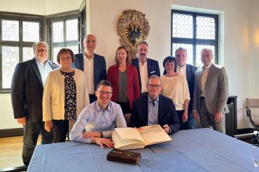 Michael Müller Trägt sich ins Goldene Buch der Stadt Nördlingen ein, Er sitzt an einem Tisch der eine Blaue Tischdecke drüber hat. Hinter ihn sind Personen.