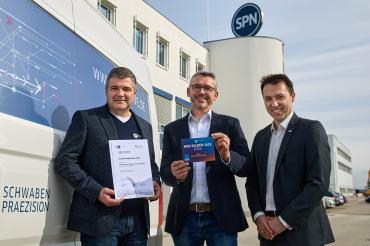 Jürgen Sieber (Verantwortlich für gewerbliche Ausbildung), Rainer Hertle (Techn. Geschäftsführer) und IHK Regionalgeschäftsführer Matthias Hausmann v.l. mit Urkunde und Aufkleber vor dem Firmengebäude von SPN, Schwaben Präzision in Nördlingen