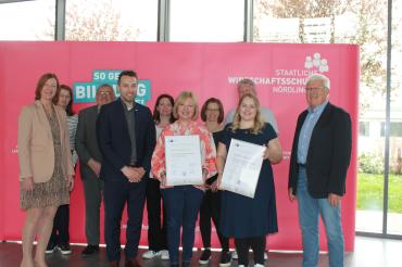 Menschen die vor einer werbewand stehen und zwei Zertifikate in der Hand halten