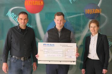 Ein Mann, der einen großen Check der hält und in der Mitte steht, neben ihm steht links und rechts eine Person