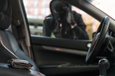 Symbolbild eines maskierten Mannes an einem Auto. Im Vordergrund ein Geldbeutel. 