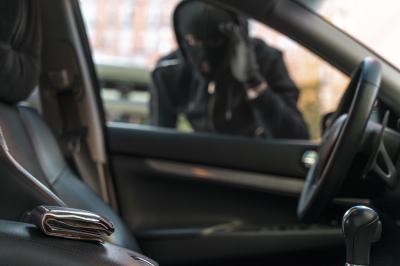 Symbolbild eines maskierten Mannes an einem Auto. Im Vordergrund ein Geldbeutel. 