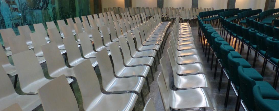 Auf dem Bild zu sehen sind mehrere Stuhlreihen in einer Veranstaltungshalle. 