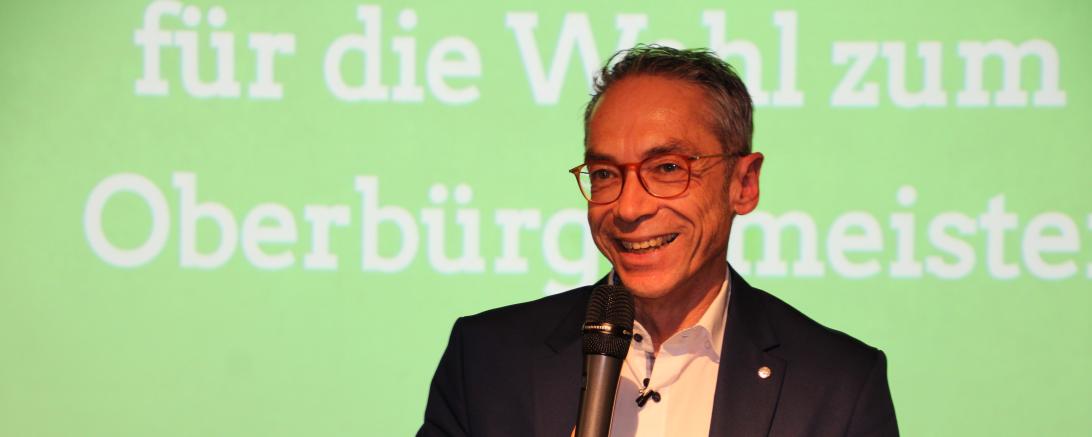 Wolfgang Goschenhofer mit Mikrofon vor einer angestrahlten Leinwand