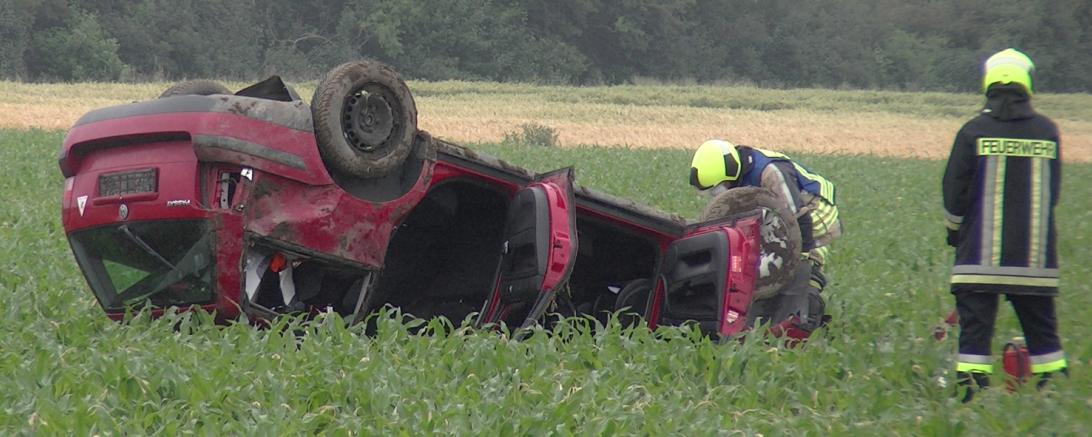 Roter Pkw liegt auf dem Dach in einem Feld, dabei zwei Feuerwehrleute