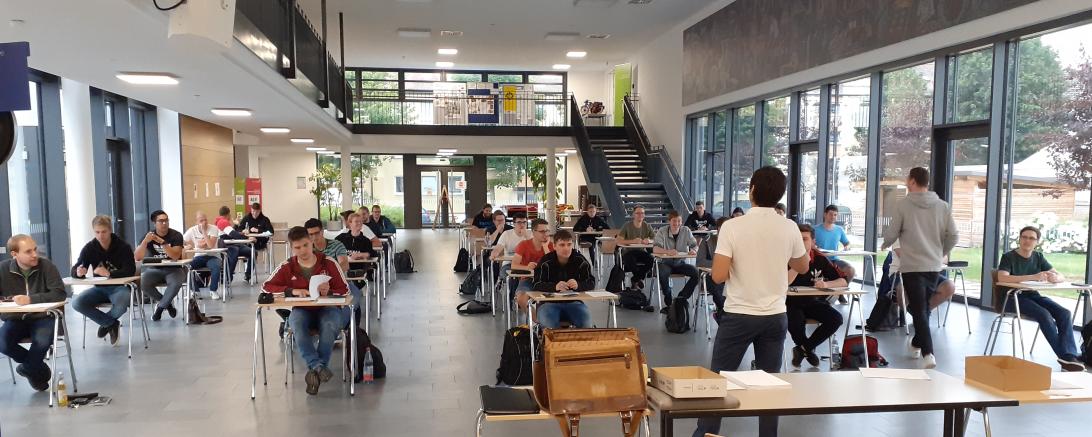 Schüler sitzen an Einzeltischen in Aula, davor eine Lehrkraft