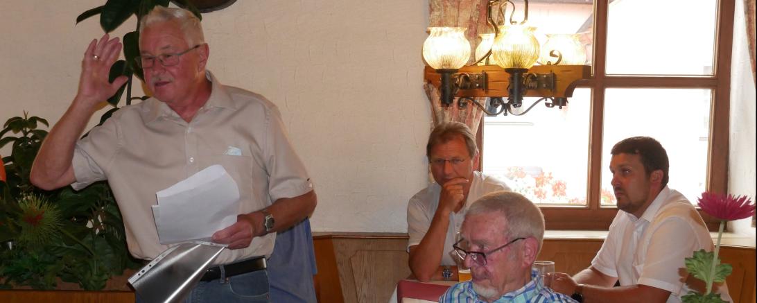 Vier Männer im Wirtshaus, drei sitzend, einer stehend, eine Rede haltend