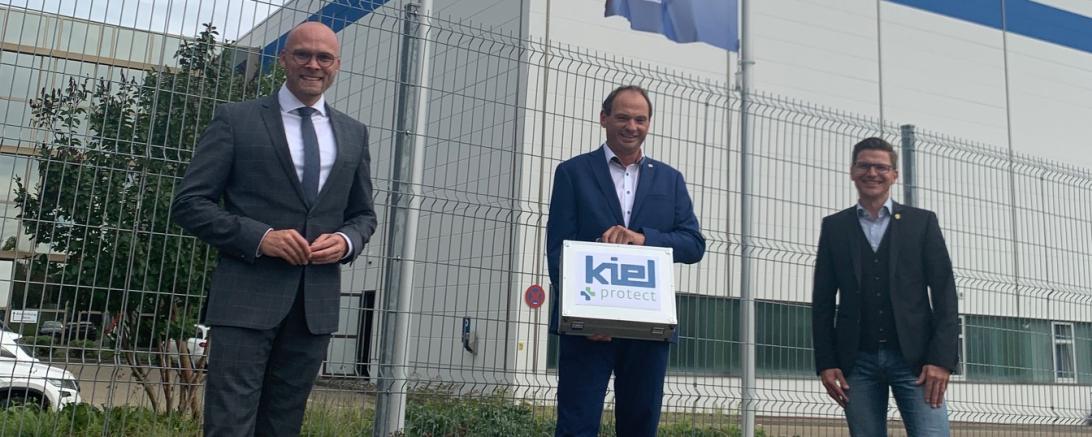 Drei Männer vor zwei Flaggen, dahinter Firmengebäude