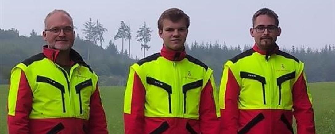 Drei Männer mit gelb-roter Warnkleidung auf Wiese vor Wald