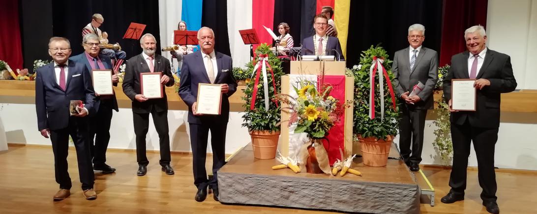 Sieben Herren im Anzug mit Urkunden vor Bühne, dahinter Flaggen