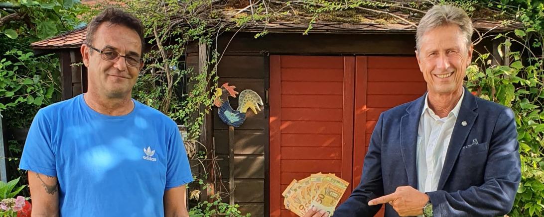 Zwei Männer vor Gartenschuppen, einer zeigt auf Geld in seiner Hand