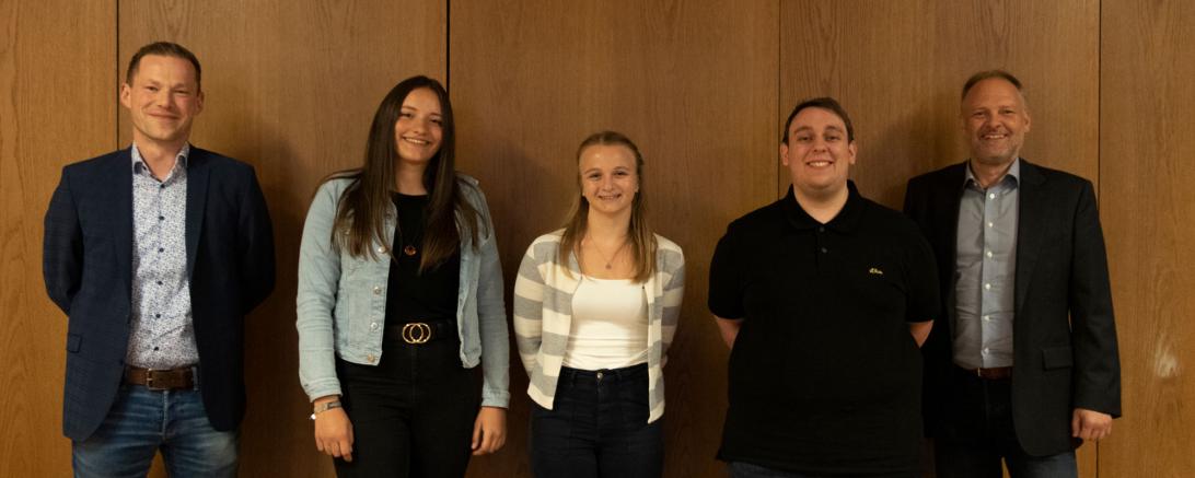 Zwei Frauen und drei Männer stehen vor einer Holzwand