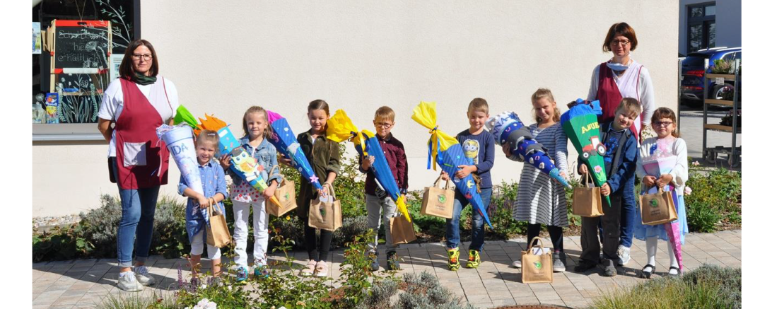 Das Bild zeigt Marion Dopfer (links) und Monika Schuster (rechts) zusammen mit den beschenkten Erstklässlerinnen und Erstklässlern vor dem Dorfladen in Rögling.