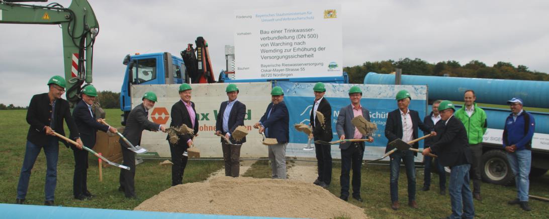 13 Herren beim Spatenstich, davor Wasserleitungsrohr