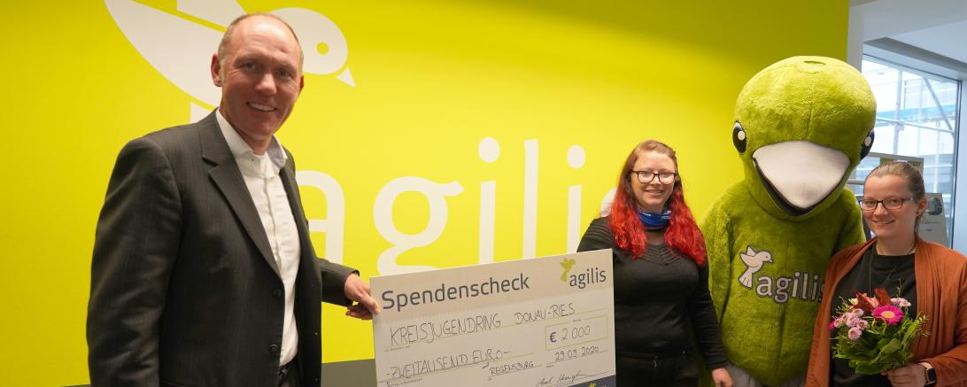 Drei Personen und ein Maskottchen mit Scheck vor agilis-Logo