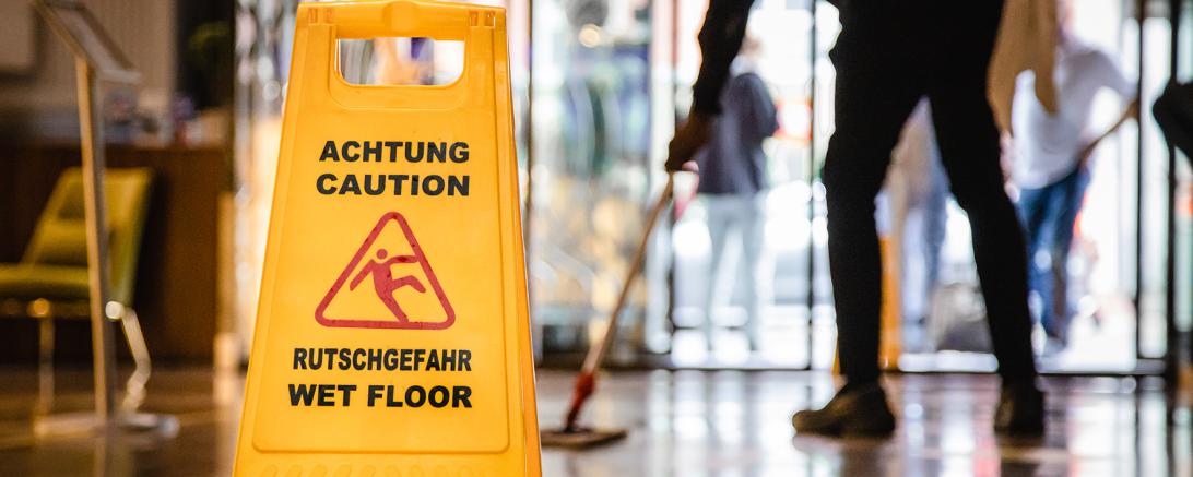 Schild Vorsicht Rutschgefahr, dahinter Gebäudereiniger beim Bodenwischen